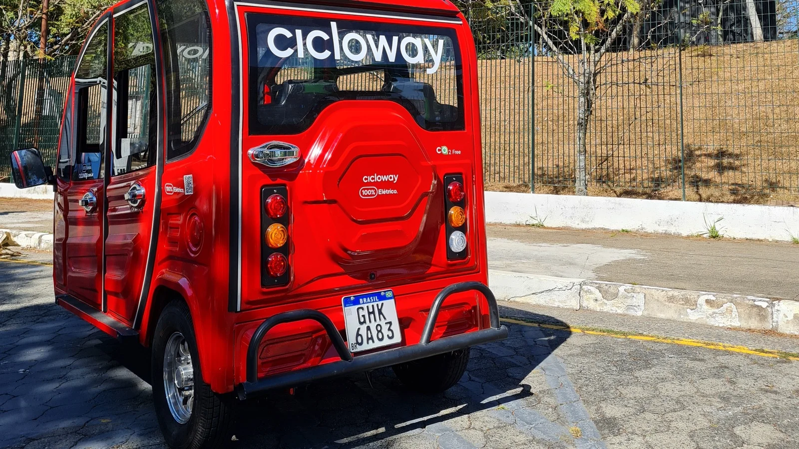 Triciclo elétrico vermelho da Cicloway visto por trás, com a placa em destaque, estacionado em rua urbana