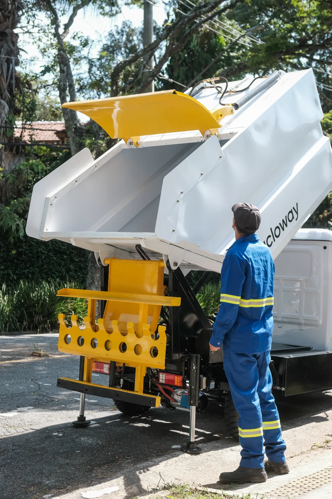 Clique para editar a descrição da imagem Operador uniformizado acompanha o baú branco de um triciclo elétrico de coleta da Cicloway basculando.